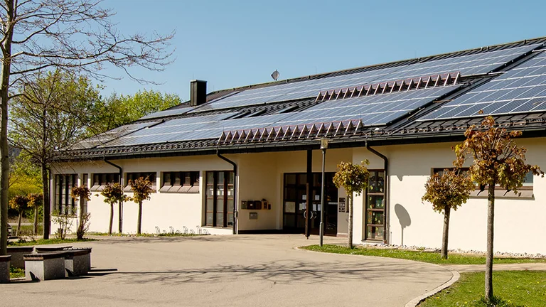 Kindergarten Oberdorf in Waltenhofen – helles Gebäude mit großflächigen Solarpaneelen auf dem Dach, gepflastertem Hof und jungen Bäumen bei sonnigem Wetter.