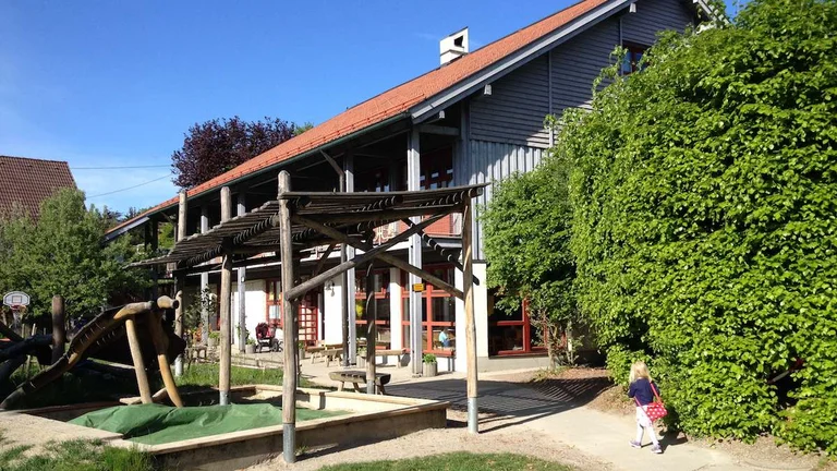 Kindergarten Hegge in Waltenhofen – zweistöckiges Gebäude mit rotem Ziegeldach, Holzpergola und Spielbereich mit Sandkasten und Klettergerüst im Garten.